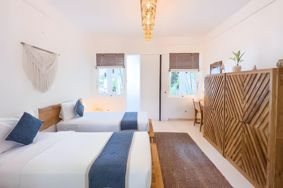 Quarto moderno com duas camas, uma cômoda de madeira e uma janela grande.