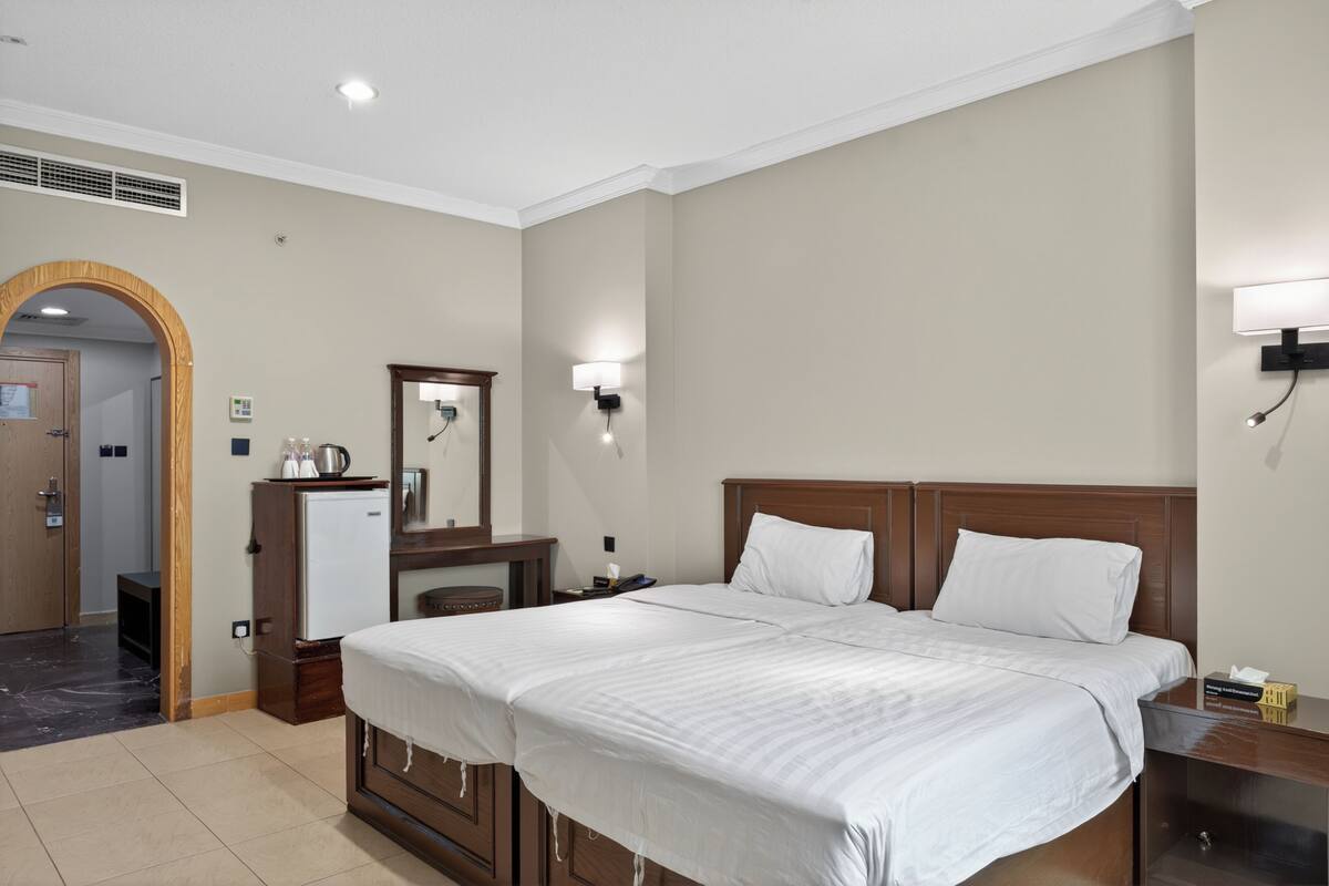 A hotel room with a large bed, a wooden headboard, a nightstand, a desk with a lamp, a mirror, and a small refrigerator.