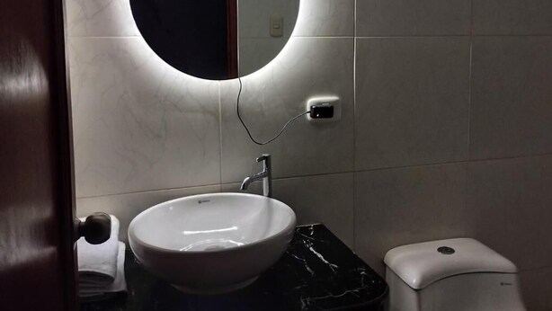 A bathroom with a round mirror, a white sink, a black countertop, and a toilet. - Alpha (Cajamarca)