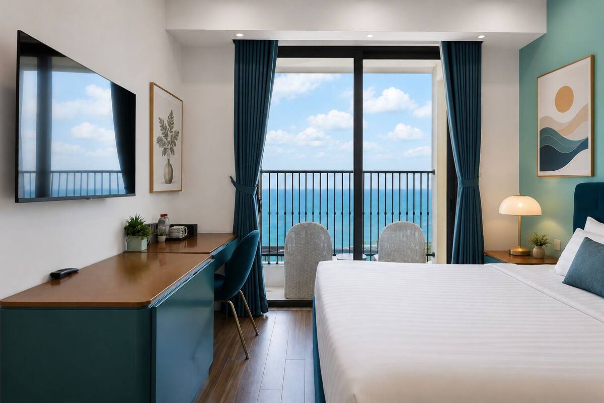Habitación de hotel con cama, escritorio, sillas, televisor y balcón con vista al mar.