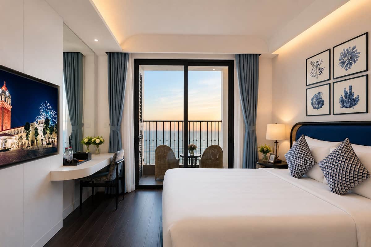 Habitación de hotel con cama, escritorio, silla, balcón con cortinas y cuadros en la pared.