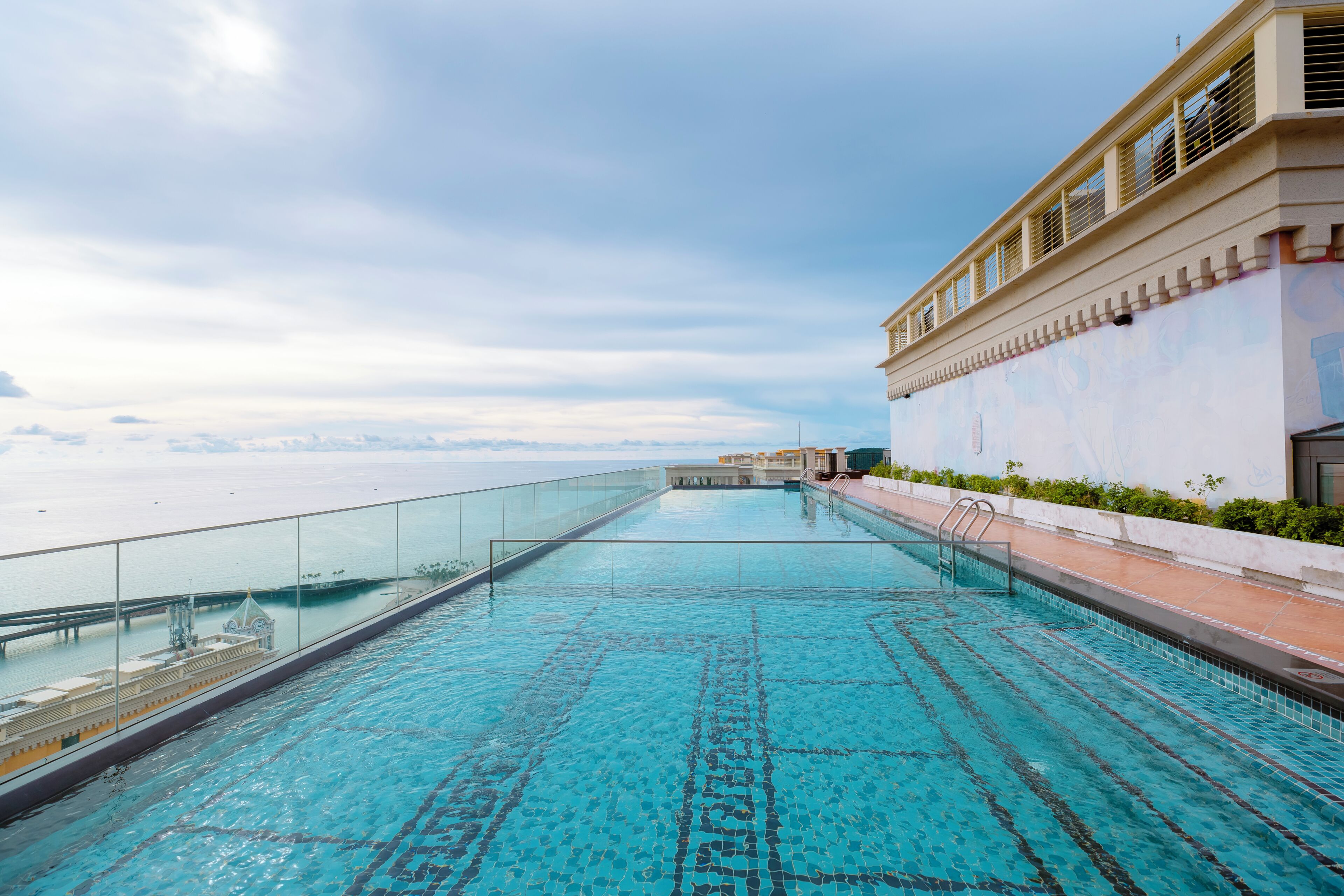 Una piscina infinita en la azotea con barandilla de cristal, con vistas a una masa de agua y un paisaje urbano lejano.