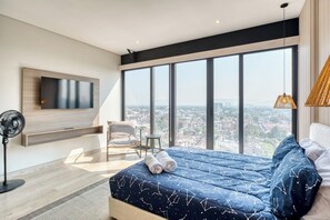 Ein modernes Schlafzimmer mit einem großen Bett, einem an der Wand montierten Fernseher, einem Sessel und Blick auf die Stadt durch große Fenster.