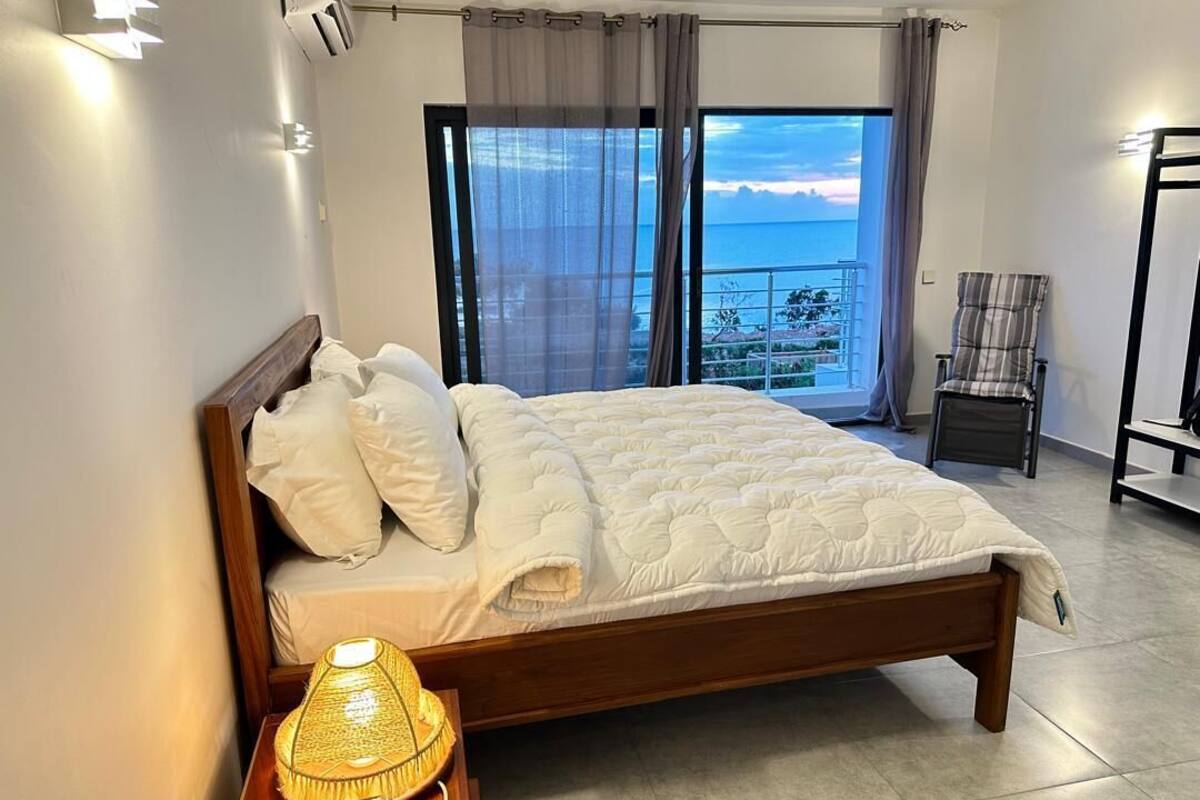 A bedroom with a bed, a nightstand with a lamp, a chair, and a view of the sea through a balcony.