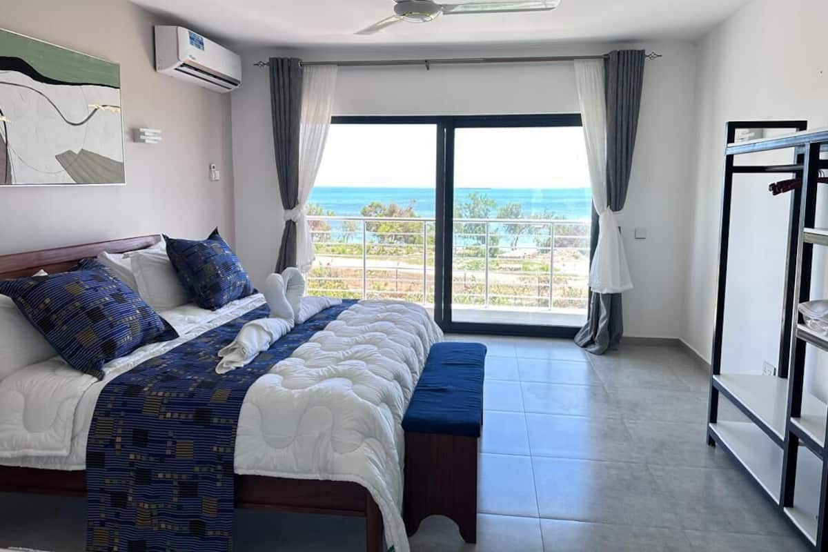 A bedroom with a bed, a bench, a ceiling fan, an air conditioning unit, a view of the ocean, and a balcony.
