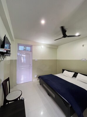 Una habitación con una cama, un ventilador de techo, un televisor en un estante, una puerta y una silla.