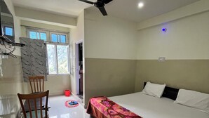 A bedroom with a bed, a chair, a ceiling fan, and a window. - Hotel Manimahesh Bharmour (Brahmaur)