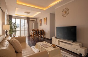 Ein modernes Wohnzimmer mit Sofa, Fernseher, Essbereich und großen Fenstern.
