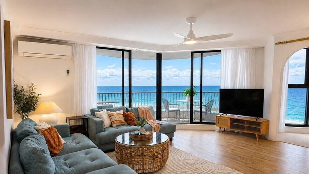 Een moderne woonkamer met een grote bank, een salontafel en een televisie. Er is een balkon met uitzicht op de oceaan.