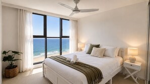 Een slaapkamer met een groot bed, een plafondventilator, een nachtkastje met lamp, een kamerplant en uitzicht op de oceaan.