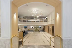 Eine Hotelhalle mit einem zentralen Kronleuchter, einer Rezeption und einer Treppe zu einer oberen Ebene.