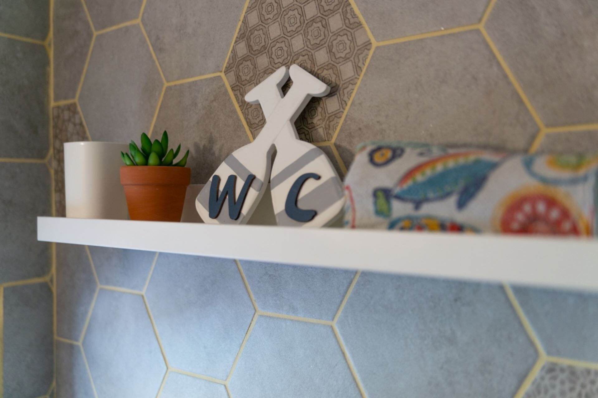 A bathroom shelf with a potted plant, a towel rack with letters, and a colorful pillow.