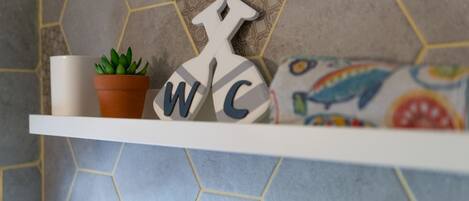 A bathroom shelf with a potted plant, a towel rack with letters, and a colorful pillow.