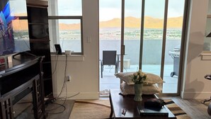 Ein Wohnzimmer mit Fernseher, Kamin, Essbereich mit Tisch und Stühlen sowie eine große Glastür, die auf einen Balkon mit Blick auf das Wasser und die Berge führt.
