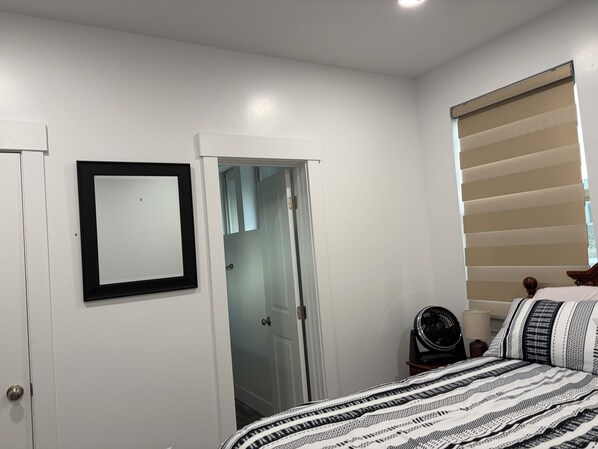 A bedroom with a bed, a bedside table with a lamp, a fan, a window with blinds, and a door leading to a bathroom.