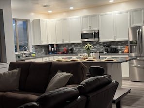 A modern kitchen with white cabinets, stainless steel appliances, and a dining area with a brown leather couch.