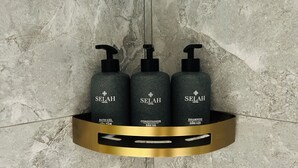 Drei Flaschen Badesets von Selah Hotel auf einem regal mit goldener Kante, vor einer strukturierten Wand.