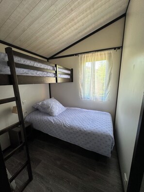 Una cama individual con escalera, una cama nido con colcha estampada, una ventana con cortinas de encaje y suelo de madera.