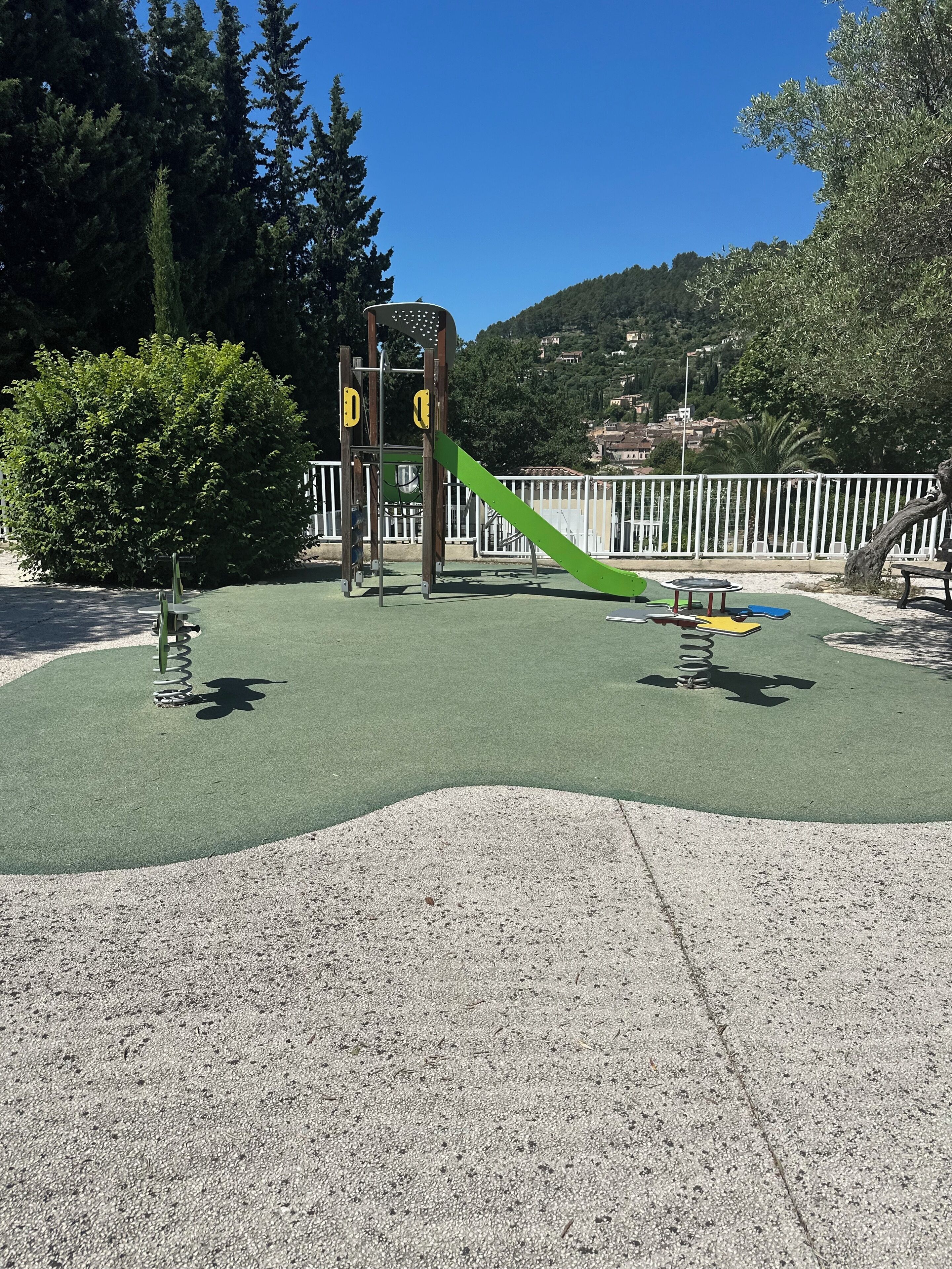 A playground with a slide and climbing structures, surrounded by trees and a white fence.