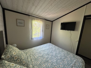 A bedroom with a bed, a window with curtains, a wall-mounted TV, and a door.