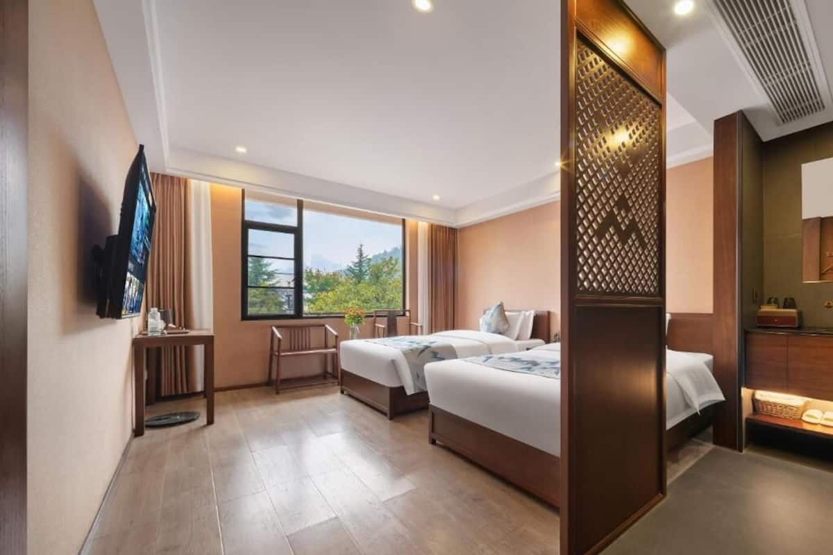 Quarto de hotel com duas camas, televisão, escrivaninha e janela com vista para o verde.