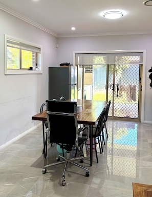 A dining area with a large table, chairs, a black office chair, a refrigerator, and a sliding glass door leading outside.