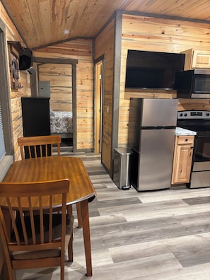 A compact kitchen with wooden walls, a dining table, chairs, a refrigerator, a microwave, and a stove.