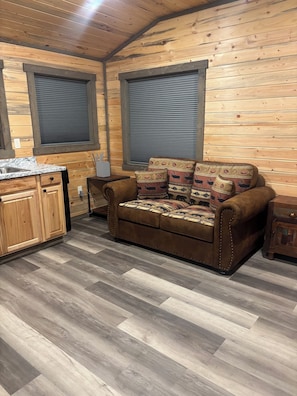 A cozy cabin interior with a wooden floor, a sofa with patterned cushions, a small wooden cabinet, and a window with blinds.