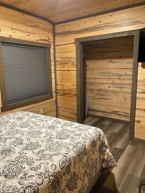 A wooden cabin room with a bed, a window with blinds, and a door leading to another room.