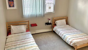 Ein Schlafzimmer mit zwei Einzelbetten, einem Fenster mit Jalousien, einer kleinen Ablage mit einer Vase und einem gerahmten Bild an der Wand.