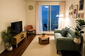 Ein Wohnzimmer mit Fernseher, einer Couch, einem Sessel, einem kleinen Tisch und einem Balkon mit Tisch und Stühlen.