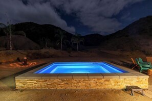 Ein rechteckiger Pool mit Steinbordstein, umgeben von einer Erdbereifung mit Stühlen und einer kleinen Feuerstelle.