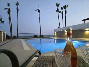 Zona junto a la piscina con sillones de descanso, una botella y vasos sobre una mesa, con vista al mar y palmeras.