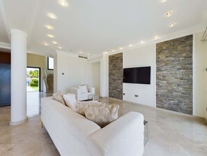 A spacious living room with a large white sofa, a flat-screen TV mounted on a stone wall, and a staircase leading to another level. - Fabulous Beach Front House (Perivolia)