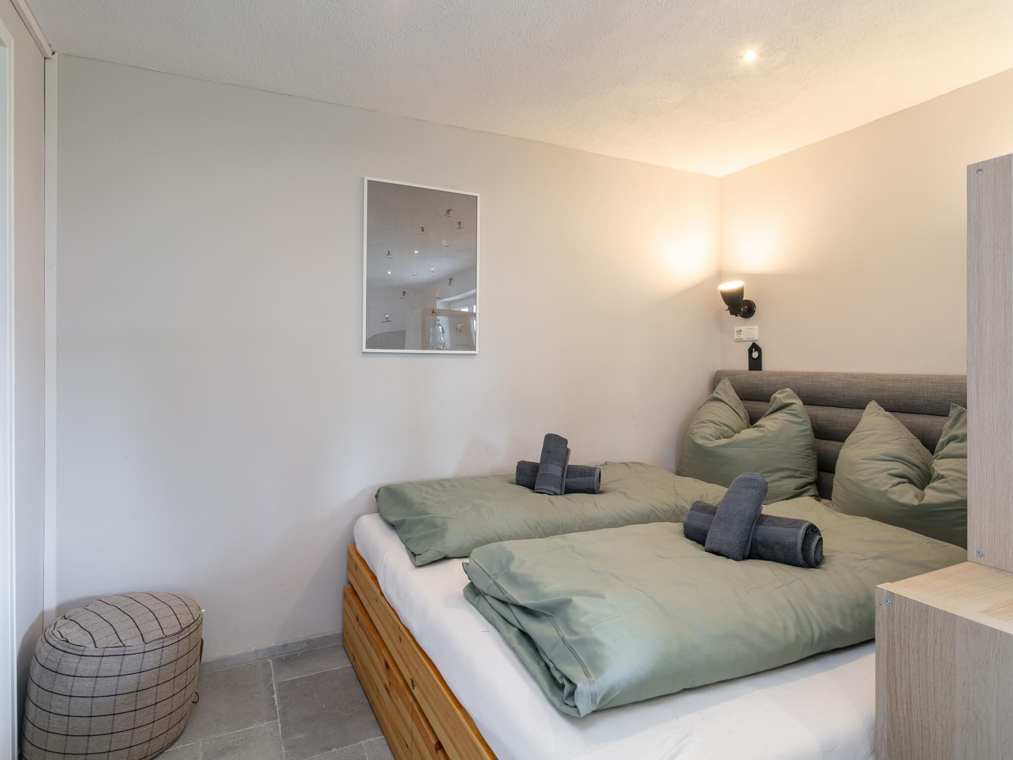 A bedroom with two beds, a wall-mounted light, a framed picture, and a small ottoman.