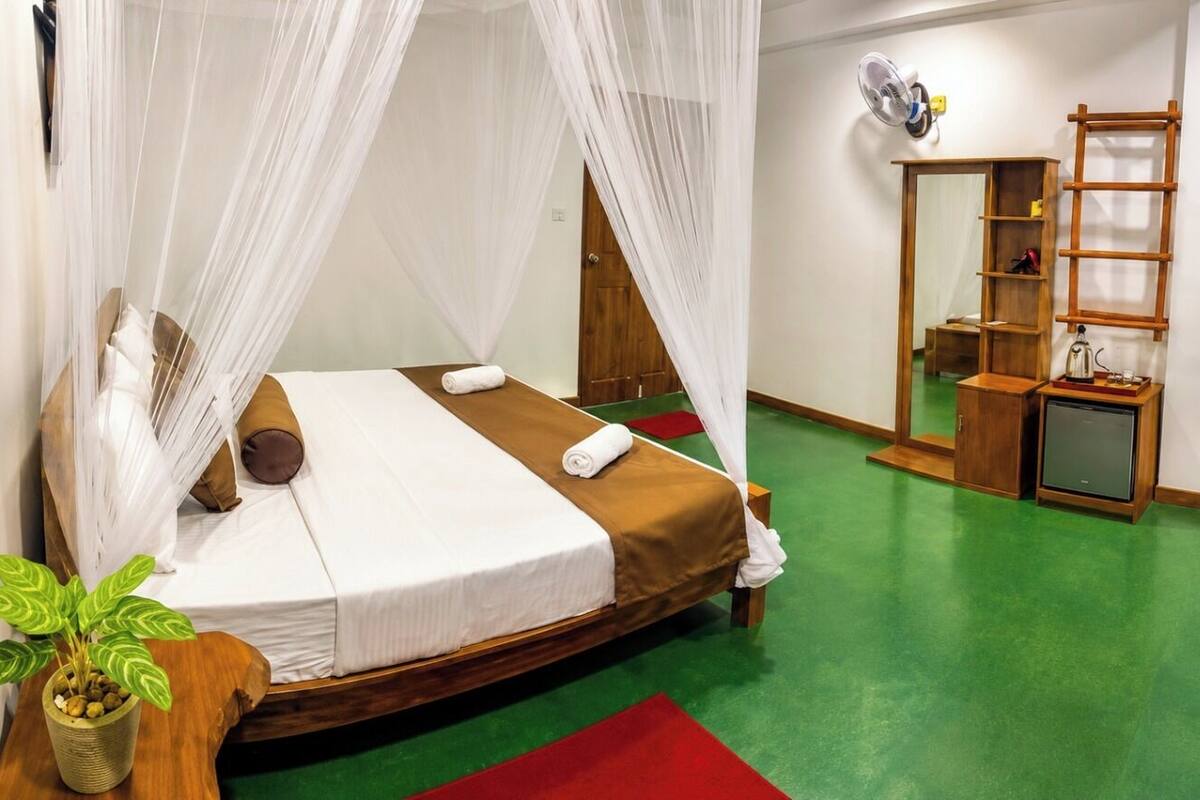 A room with a bed, a small table with a plant, a mirror, and a shelf with a fan.