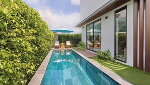 Una piscina moderna al aire libre con cubierta de madera, rodeada de exuberante vegetación y un área de patio con paredes de vidrio.