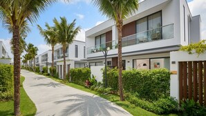 Un hotel moderno con un camino bordeado de palmeras y follaje, con balcones y un diseño limpio y contemporáneo.