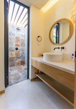 Un baño con ducha de piedra, lavamanos blanco, espejo redondo y tocador de madera.