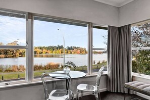 A room with a large window offering a view of a lake and autumn foliage. There is a round table with two chairs and a bench. - Lake View Apartment with parking (Hamilton)