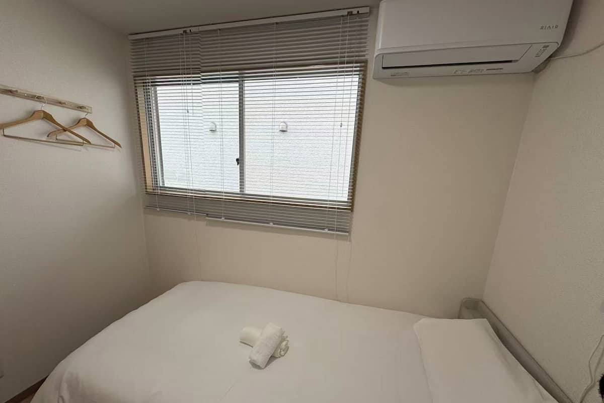 A small, neatly made bed with a white mattress and pillows, a window with blinds, a wall-mounted air conditioner, and a wooden hanger on the wall.