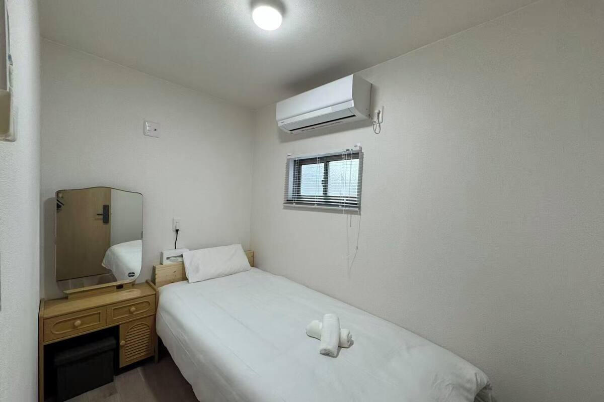 A single-bed room with a wooden dresser, a mirror, a window with blinds, an air conditioning unit, and a ceiling light.