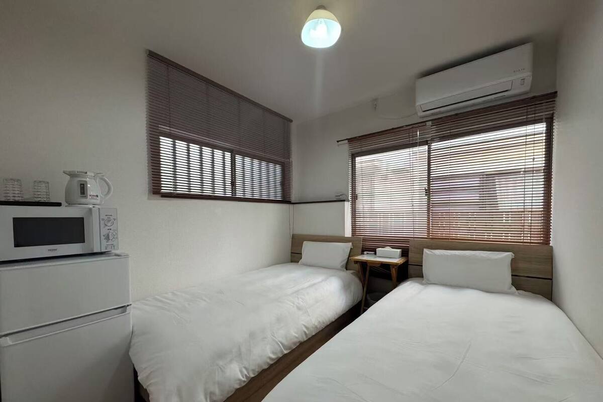 A hotel room with two single beds, a kitchenette with a microwave, and an air conditioning unit.