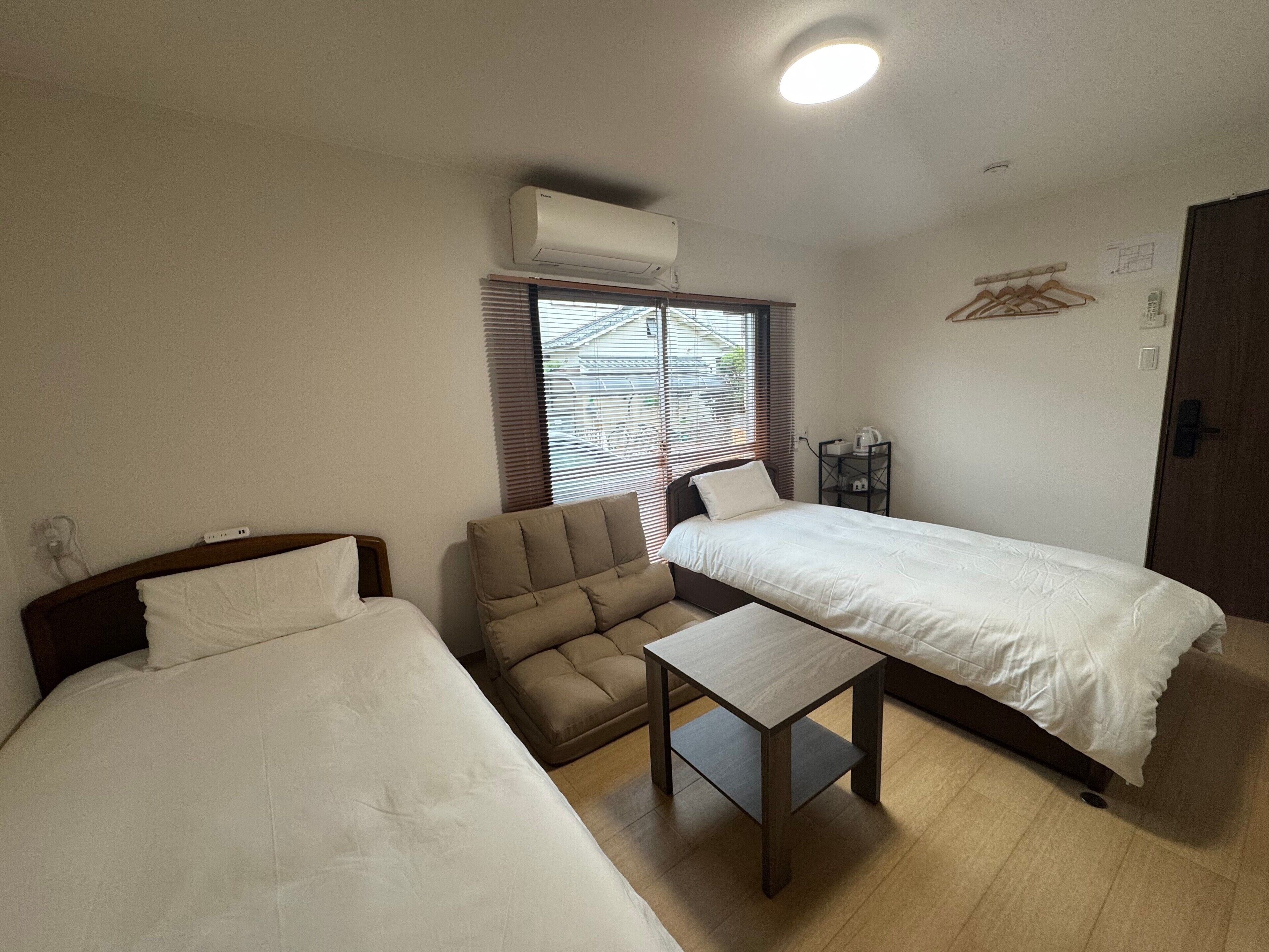 Une chambre avec deux lits, une petite table, un canapé, un appareil de climatisation et une fenêtre avec des stores.