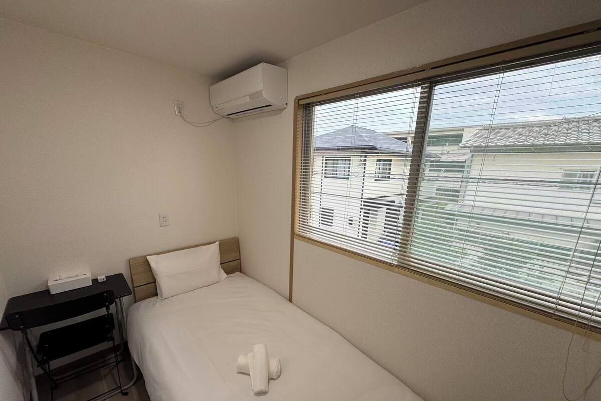 A single bed with a white mattress and pillow, a small desk with a chair, and a window with blinds.