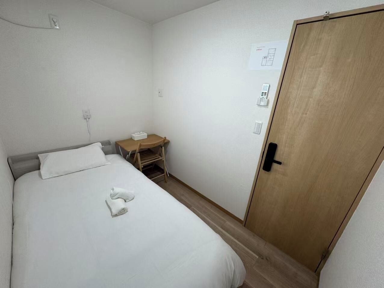 Een enkel bed met een witte deken en een opgevouwen handdoek, een houten nachtkastje met een lamp, een houten deur en een stopcontact.
