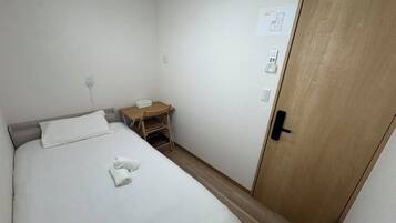 Een enkel bed met een witte deken en een opgevouwen handdoek, een houten nachtkastje met een lamp, een houten deur en een stopcontact.