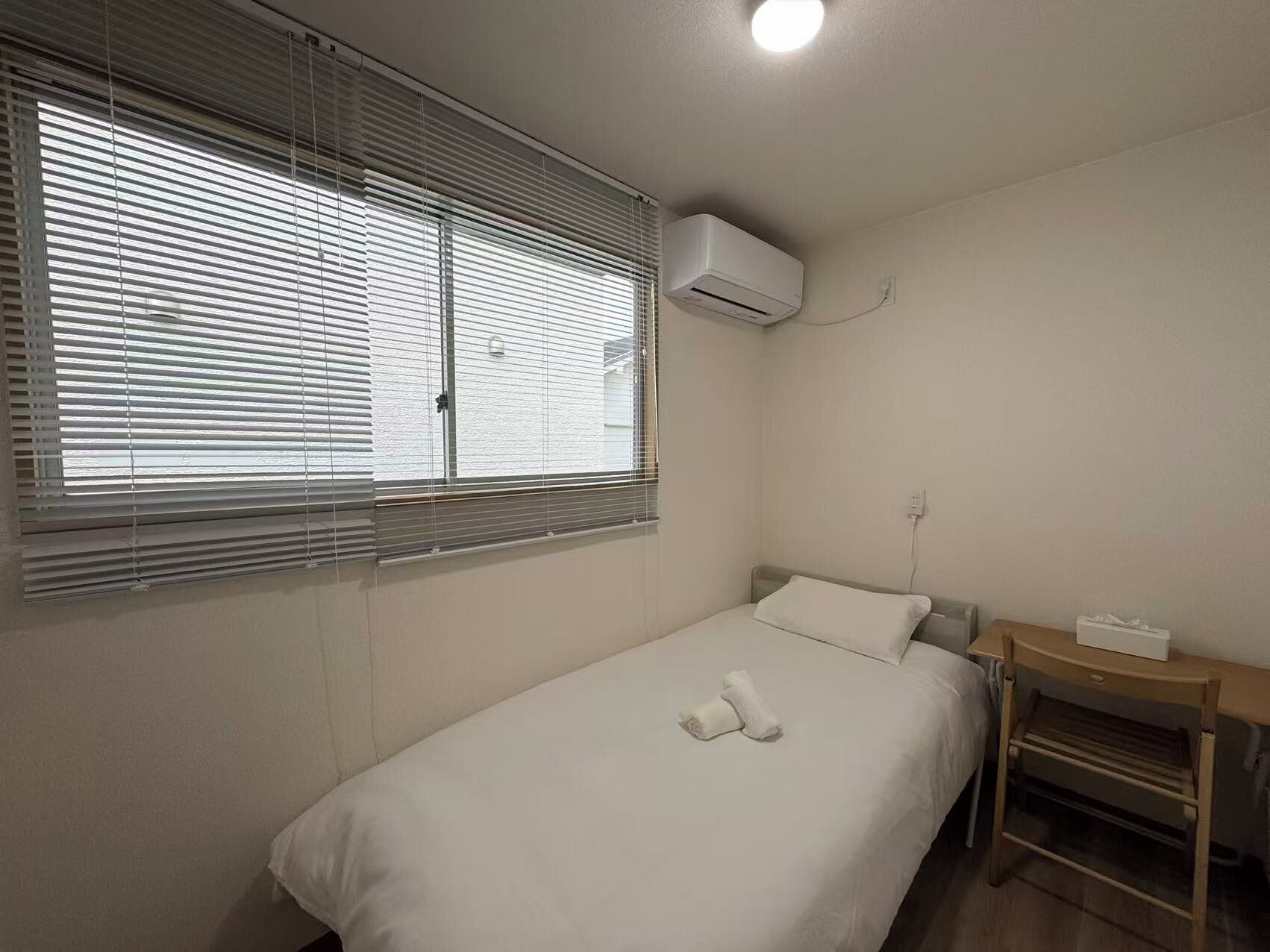 A single-bed room with a window, an air conditioning unit, a bedside table with a lamp, and a small desk with a chair.