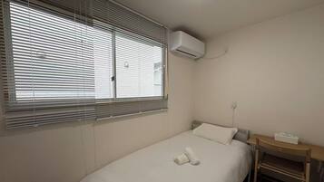 A single-bed room with a window, an air conditioning unit, a bedside table with a lamp, and a small desk with a chair.
