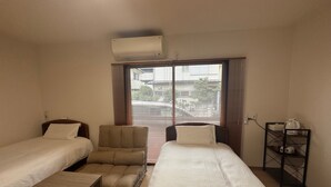 Ein Hotelzimmer mit zwei Betten, einem Sessel, einem kleinen Tisch, einem Regal mit einem Wasserkocher und einer Klimaanlage.